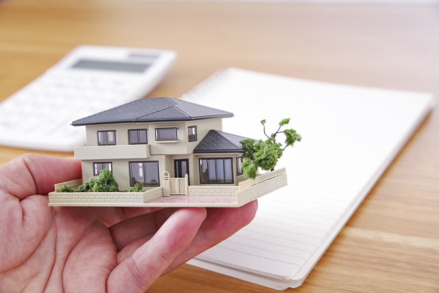 手のひらに乗せられた住宅模型と背景に置かれたノートと電卓