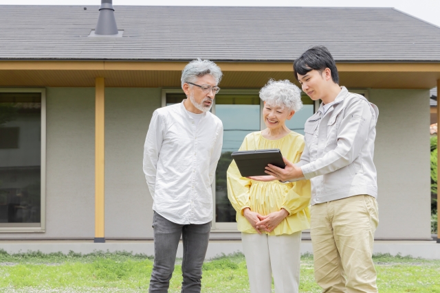 住宅の前でタブレットを見ながら相談する高齢夫婦と担当者
